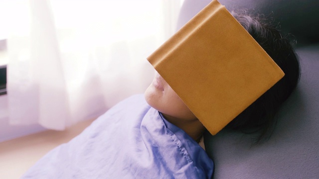 年轻的亚洲女人放松和睡着的时候，在客厅的沙发上看书，特写镜头。视频素材