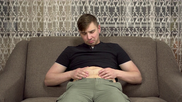 一个年轻人坐在沙发上，摸着他的胖肚子。在隔离期间体重增加视频素材