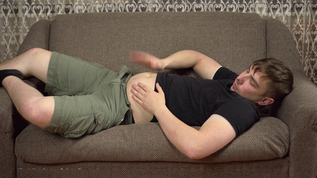 一个年轻人躺在沙发上，拍着自己的肚子。在隔离期间体重增加视频素材