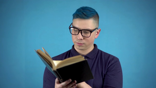 一个戴眼镜、头发是蓝色的年轻人正在读书。另一个男人手里拿着一本书，背景是蓝色的。视频素材