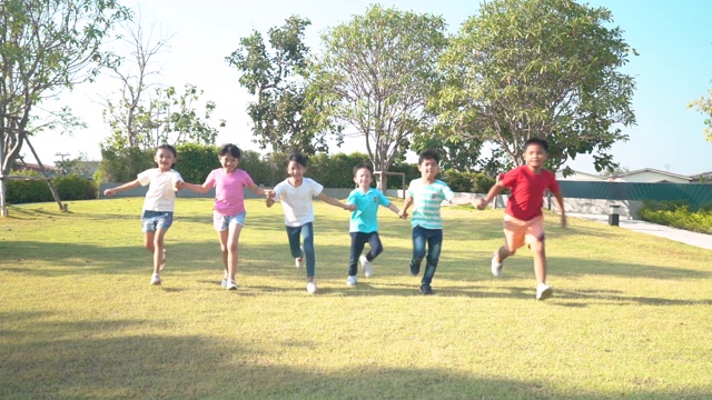 在阳光明媚的夏日，一群快乐的亚洲幼儿园小朋友穿着休闲服装，手牵着手在公园里玩耍、奔跑。视频下载