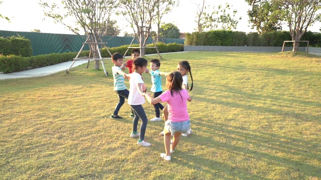 在阳光明媚的夏日里，一群快乐的亚洲微笑的幼儿园小朋友手拉手嬉戏跳舞，在公园里的绿草地上打转、站成一圈。视频下载