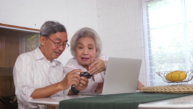 幸福的亚洲老年夫妇使用笔记本电脑和支付与信用卡在家里，家庭放松和花时间在一起在家里，退休人士和生活方式的概念视频素材