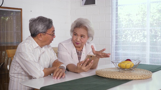 幸福的亚洲老年夫妇坐在家里使用手机，家庭放松和花时间一起在家里，退休人士和生活方式的概念视频素材