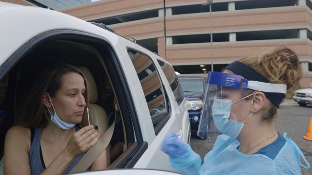 一名身穿长袍、戴着外科口罩、手套和面罩的女护士将检测棉签递给一名30多岁的女性，她坐在诊所/医院门外的COVID-19检测线上，坐在一辆白色SUV上视频下载
