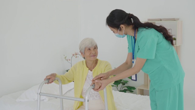 微笑的亚洲护士协助老年妇女起床。照顾护士支持病人从床上起来，并在家中走向助行器。帮助残疾老妇人站起来。视频下载