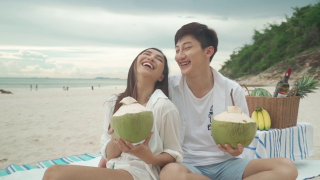 亚洲年轻夫妇坐在野餐毯上，在海滩和海边喝椰子汁，背景是热带水果。夏季、假期、假期和快乐的泰国人概念视频素材