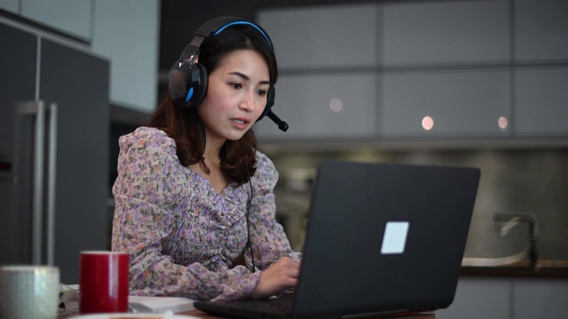 一位亚洲的中国美女在家工作，作为客户服务，用笔记本电脑和耳机在厨房打字，在线回答她的客户视频下载