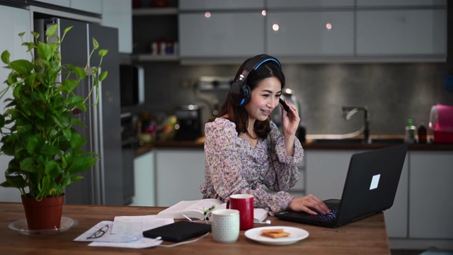 一位亚洲的中国美女在家工作，作为客户服务，用笔记本电脑和耳机在厨房打字，在线回答她的客户视频素材