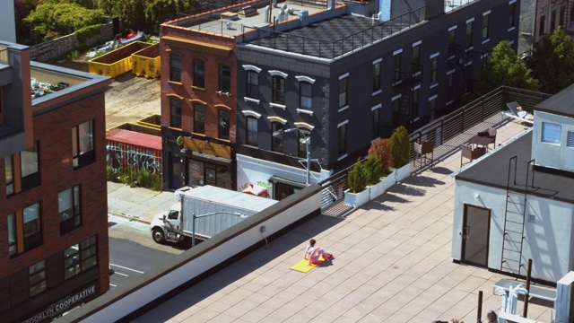一个19岁的女孩在屋顶上做着健身运动，在远处俯瞰曼哈顿——她坐着，伸展她的背部和双手。视频下载