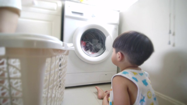 亚洲男婴坐在地板上，看着家里自动洗衣机里的衣服视频下载