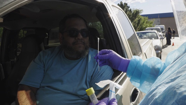 在医疗诊所/医院外的COVID-19(冠状病毒)检测队列中，一名身穿长袍、戴着外科口罩、手套和面罩的女护士向一名坐在卡车里的有色人种男性解释棉签检测视频素材