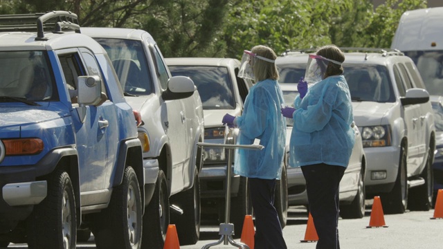在医疗诊所/医院外的COVID-19(冠状病毒)检测队列中，两名身穿长袍、戴着外科口罩、手套和面罩的女护士在车内与病人交谈(第二波)视频素材