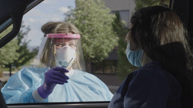 一名身穿长袍、戴着外科口罩、手套和面罩的女护士走向坐在车内的女性患者，在医疗诊所/医院外的COVID-19(冠状病毒)检测队列中解释棉签检测视频素材