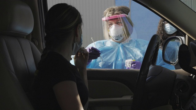 在医疗诊所外的COVID-19(冠状病毒)检测队列中，一名身穿长袍、戴着外科口罩、手套和面罩的女护士走向一名坐在车内的女性患者，用棉签擦拭她的鼻子视频素材