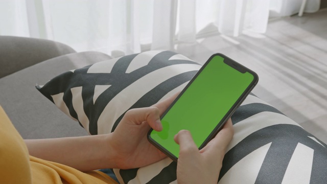 一名女子坐在家中的沙发上，手里拿着智能手机的绿色屏幕模型，上面写着信息。视频下载