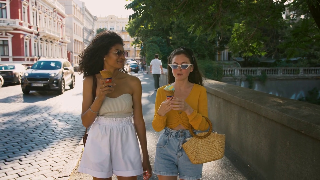 两个年轻的女孩穿着休闲装，戴着太阳镜，一边吃着冰淇淋，一边微笑着聊天，走在城市的街道上。慢动作视频下载