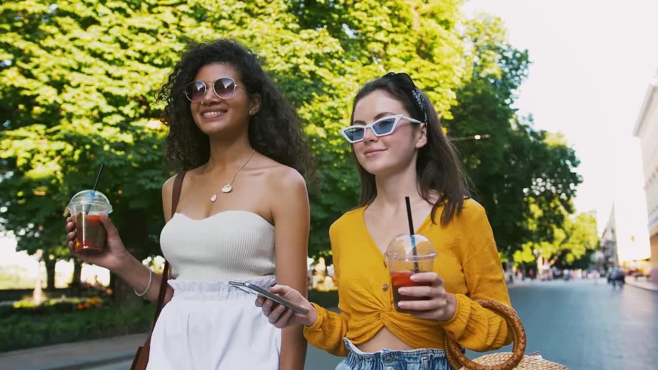 女孩们用塑料杯端着冷茶。笑着，看着手机上的视频，看起来很惊讶。沿着城市大道散步。特写，慢镜头视频素材