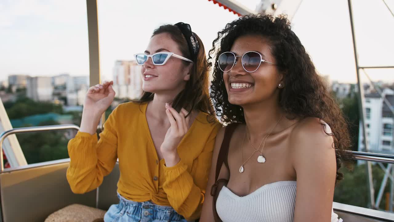 年轻的女孩们戴着太阳镜，穿着休闲装，坐在摩天轮的亭子里，微笑着，交谈着，看起来很惊讶。慢镜头特写,视频素材