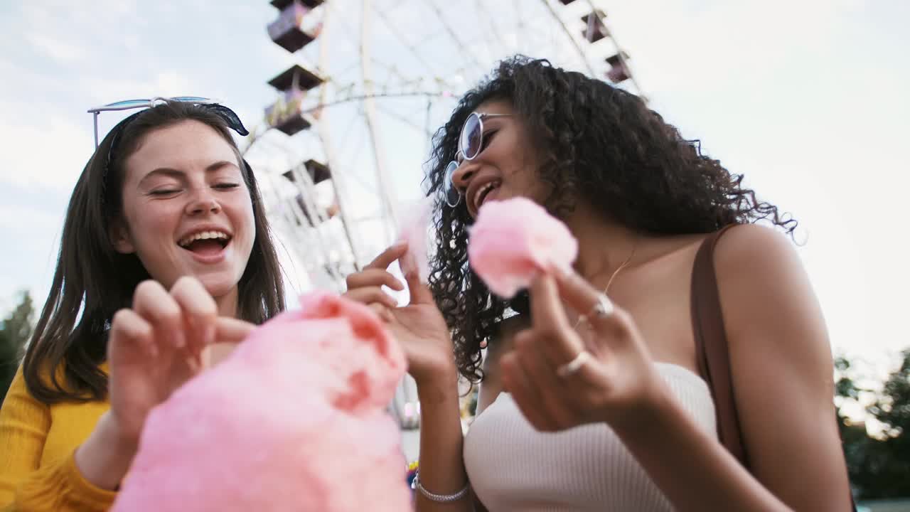 在公园里，面对摩天轮和蓝天，两位年轻的女性一边吃着粉色的棉花糖，一边微笑着摆姿势。慢动作，特写视频素材
