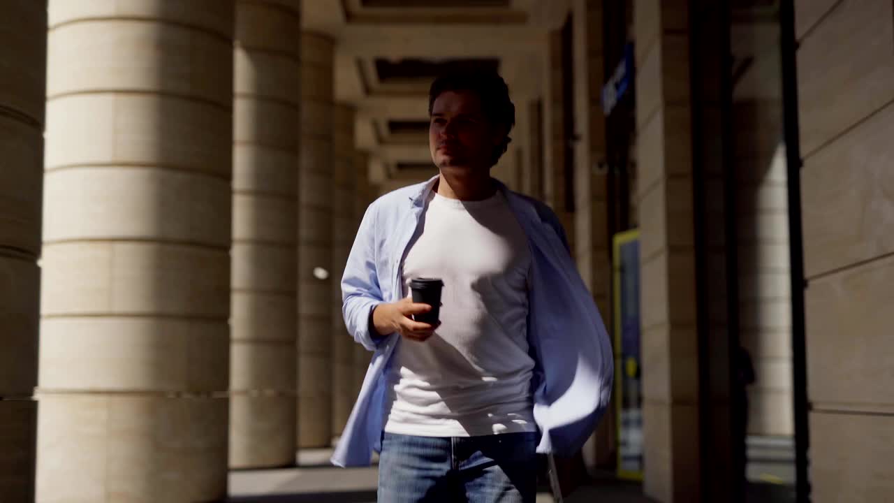 中镜头的正面，自信的年轻企业家走在拱廊下的工作，带着外卖咖啡杯和手提电脑视频下载