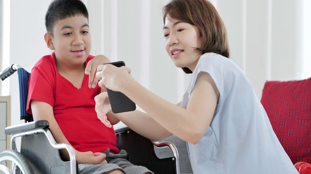 10-11岁的亚洲残疾男孩坐在轮椅上，与母亲或看护一起在家通过智能手机学习在线课程。2019年残疾人募捐视频素材