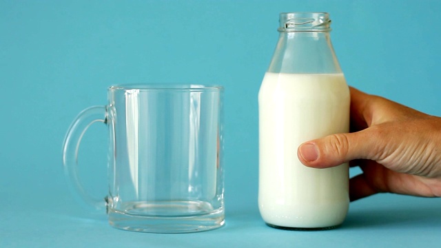 女性手拿牛奶瓶，将牛奶倒入蓝色背景的玻璃杯中，正面。塑料免费视频下载