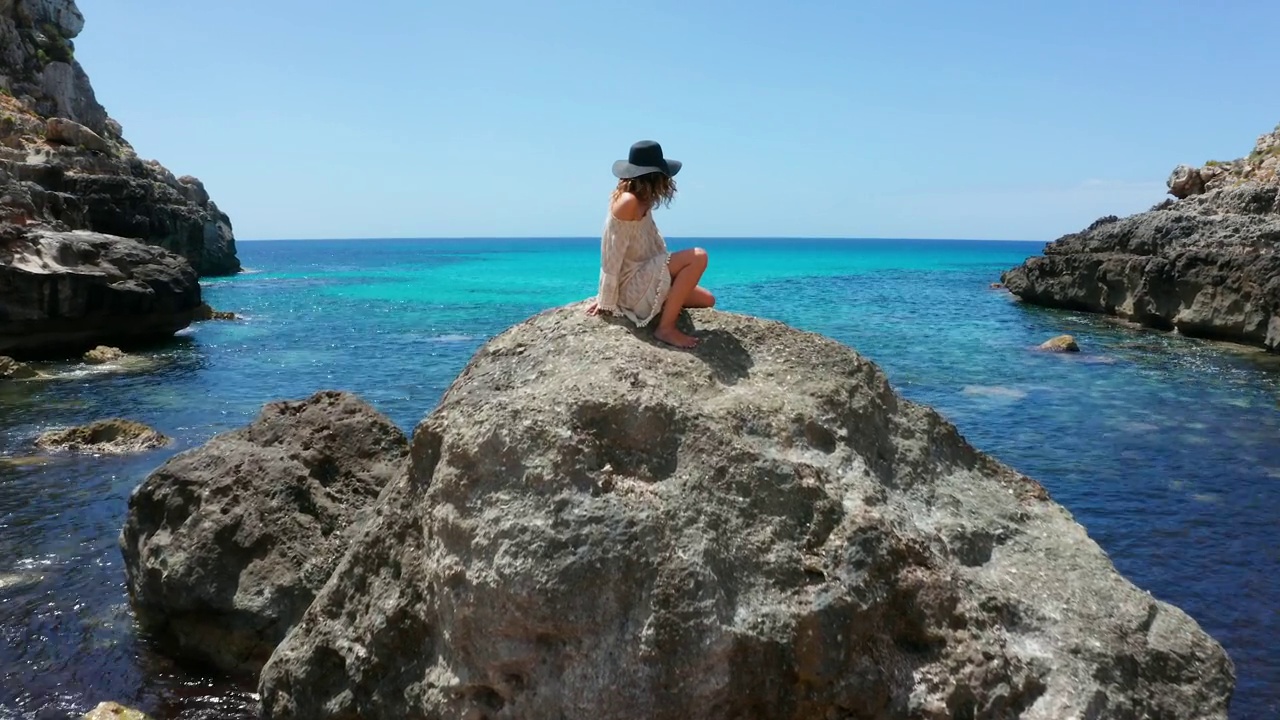 无人机在地中海上空拍摄的航拍画面，中间有一块岩石。岩石上方，一位戴着帽子坐在顶上的美丽女人。4 k视频下载