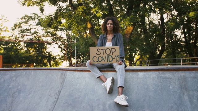 一位穿着休闲服的美国黑人女孩坐在滑板场的水泵道上，展示写有“阻止Covid-19”字样的硬纸板海报。慢动作视频下载