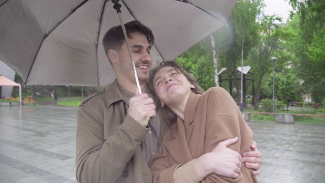 英俊的年轻人亲吻幸福的爱的女人把她的头靠在他的肩膀上。特写白种人幸福的夫妇约会在城市街道在雨天阴天。视频下载