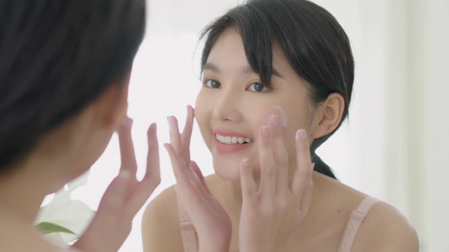美丽的年轻亚洲女人微笑看着镜子检查脸和皮肤护理霜和化妆品为年轻化和卫生，美丽的女孩快乐清洁面部与乳液和健康。视频素材
