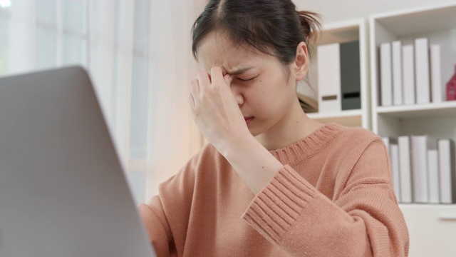 疲惫的年轻亚洲女性使用笔记本电脑，在长时间使用电脑工作后，在家庭办公室感觉眼睛疲劳。视频素材