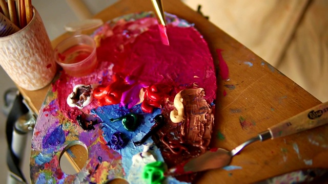女艺术家用厚厚的画笔在调色板上混合不同颜色的油粉色颜料。近距离观察女性的手混合颜色。五彩缤纷的调色板。高角度视频下载