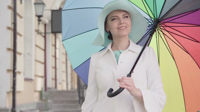 镜头靠近优雅的成年白人妇女站在户外，带着五颜六色的伞和微笑。肖像迷人的美丽的中年女士享受晴朗的一天在城市。视频素材