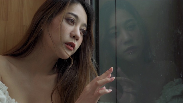 4K沮丧年轻美丽的亚洲女人独自住在公寓的窗户旁。孤独悲伤的女孩坐着，望着窗外的城市，思考着生活中的问题。负面情绪的概念。视频素材