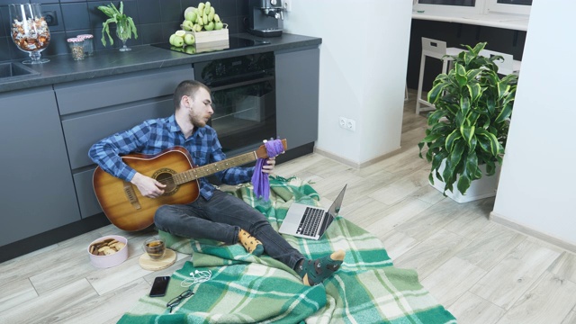 一位专业吉他手坐在家里厨房的地板上调吉他。隔离期间，音乐家在家弹奏吉他。在线吉他课程视频下载