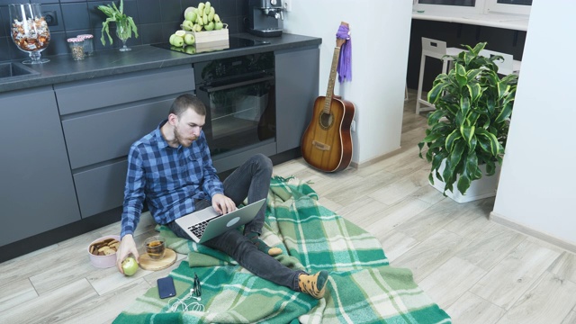 年轻的大胡子男子在家里使用笔记本电脑进行远程项目工作。男性使用电脑，坐在厨房地板上远程工作，吃苹果，玩得开心。在隔离区在家工作视频下载