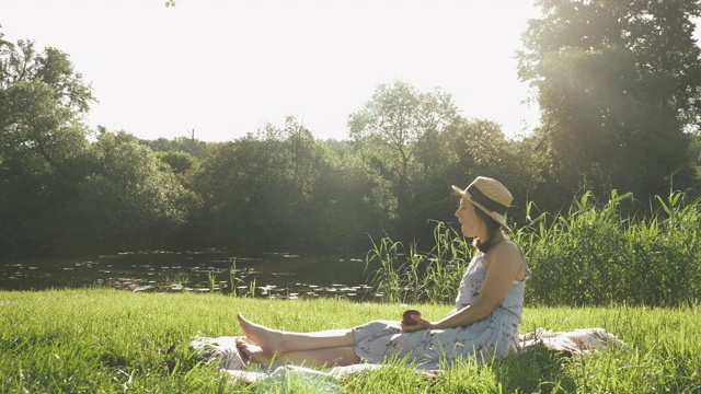 放松的年轻女子享受阳光周末的暑假在公园周围的树木和河流。戴着帽子，穿着衣服，坐在河边的草地上，吃着苹果，放松着视频素材