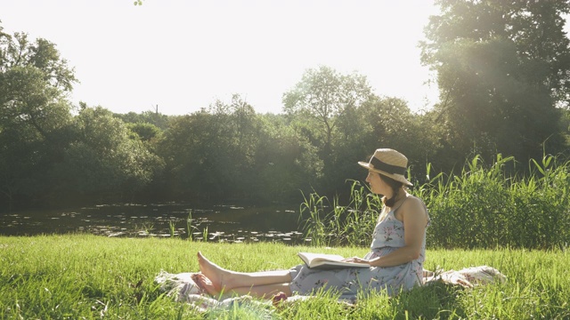 宁静的年轻女子在河边的公园里享受着温暖的阳光。可爱时尚的女性坐在毯子上，在河边的草地上看书日记。在夏日周末呼吸新鲜空气的女士视频素材