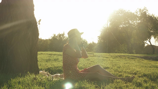 年轻女子戴着复古草帽和连衣裙，坐在公园的格子上，戴着耳机，听着音乐，享受着美丽的夏日日落。女性在公园里坐着时会戴上耳机视频素材