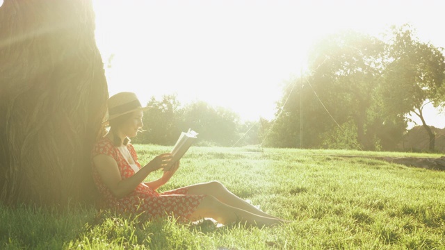 可爱的年轻女子休息在公园里，在阳光明媚的夏天读书。女孩倚在一棵大树上，坐在公园的草地上休息。女学生在公园看书日记视频素材
