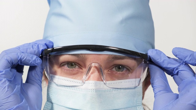 医生戴着透明的塑料眼镜，戴着医用防护口罩和蓝色的纺织品帽，看着摄像机，扶直了他的眼镜视频下载