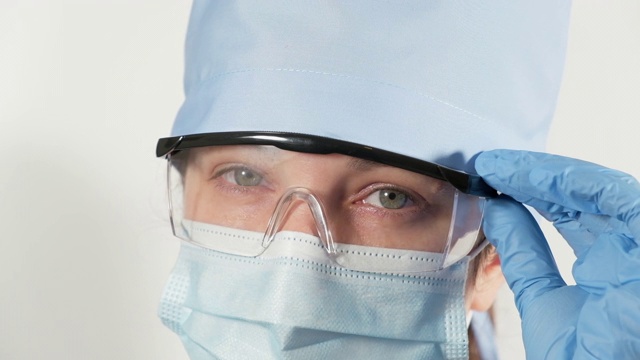 一名戴着一次性医用口罩、眼镜和纺织品帽的妇女在白色背景下看着摄像机视频下载
