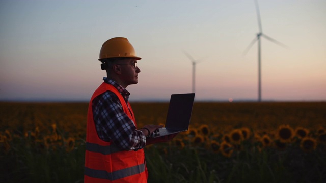 夕阳西下时，一位工程师一边在一台笔记本电脑上工作，一边调整风力涡轮机。视频素材