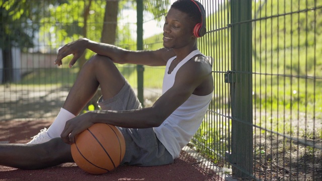 侧面的积极的非洲裔美国运动员坐在篮球场上与球和听音乐耳机。一个英俊的年轻人在阳光明媚的夏日享受假期。体育的概念。视频素材