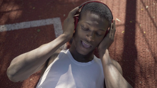 正面的运动员手拿耳机唱歌的肖像。俯视图年轻的非洲裔美国人躺在户外篮球场上，享受音乐。视频素材