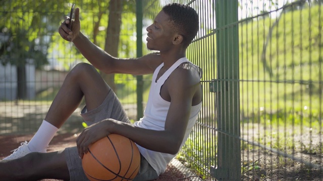 正面的非裔美国篮球运动员用智能手机自拍的侧视图。年轻人坐在球场上拿着球，面带微笑。爱好，运动，生活方式，运动精神。视频素材