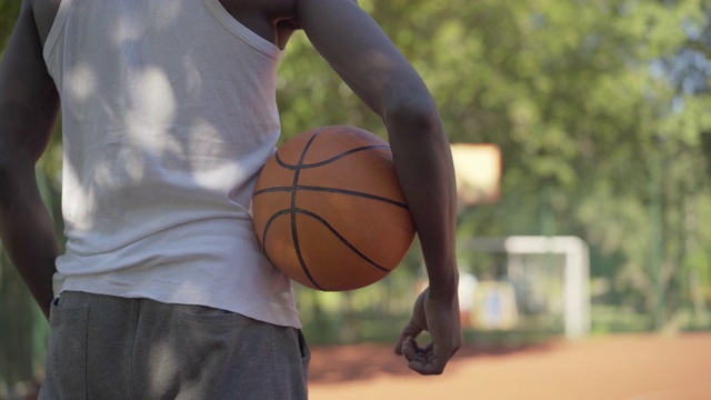 在篮球场上，一个不认识的运动员站着一个橙色的球。一个年轻的非裔美国男子在阳光明媚的日子穿着运动服在户外进行运动游戏。视频素材