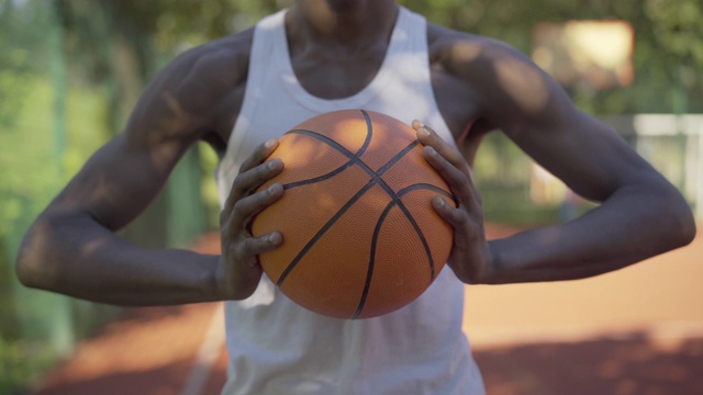 一个非裔美国运动员在室外篮球场上举着橙色的球。年轻的运动男子站在户外在阳光明媚的夏日运动器材。视频素材