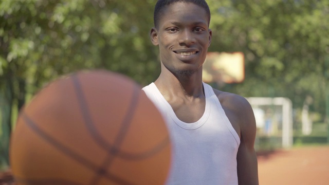 中景的非洲裔美国人伸展球相机和微笑。一个自信的年轻运动员在户外篮球场上摆姿势的肖像。快乐,体育,生活方式。视频素材